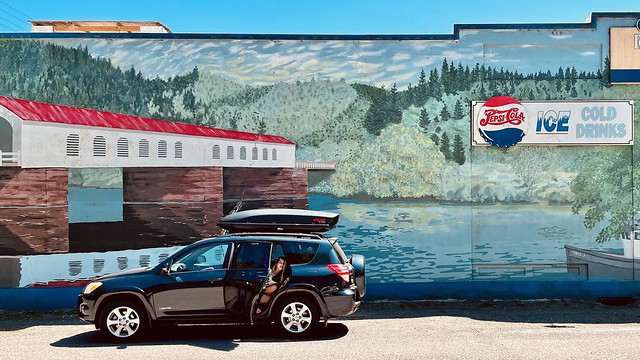 A Toyota RAV4 parked in front of building mural with covered bridge, forest landscape, and Pepsi ad