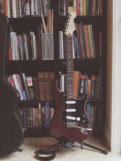 a maroon fender stratocaster electric guitar in a stand in front a tall bookshelves filled with books. An acoustic guitar case leans on the left side and drumsticks are visible on one of the shelves